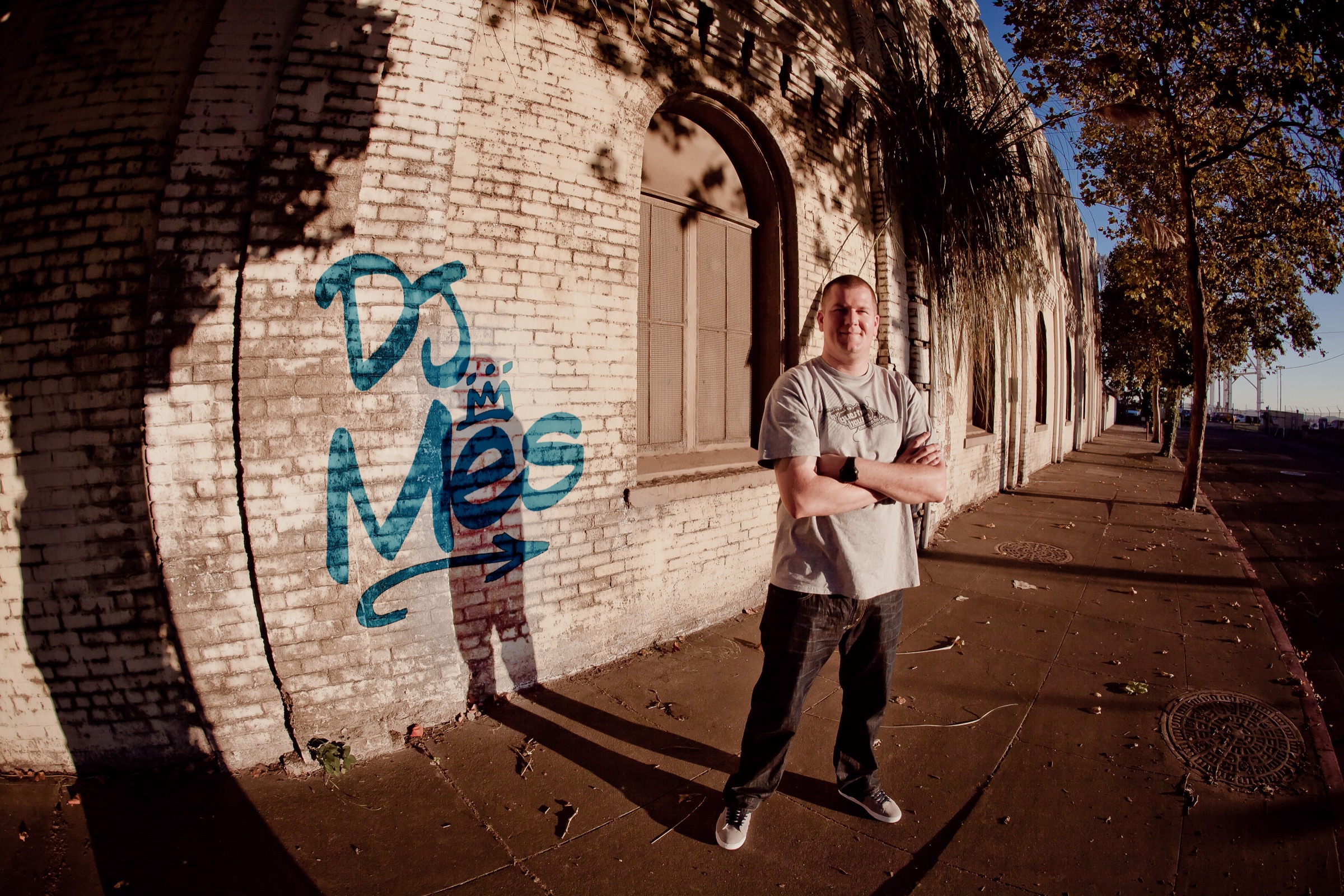 DJ Mes in front of a brick wall in Oakland with his name tagged in blue graffiti